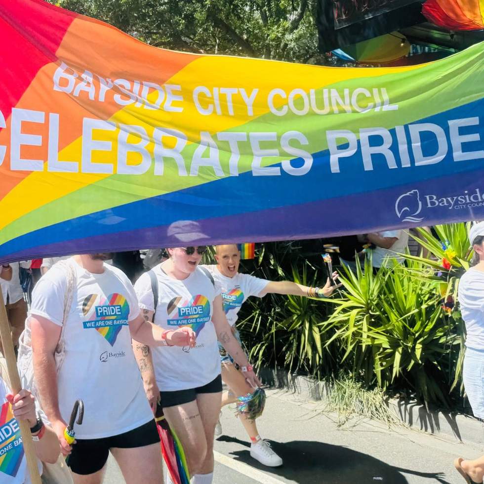 Members of staff in matching t-shirts, holding rainbow umbrellas. They are marching with a large rainbow banner that reads Bayside City Council celebrates pride. 