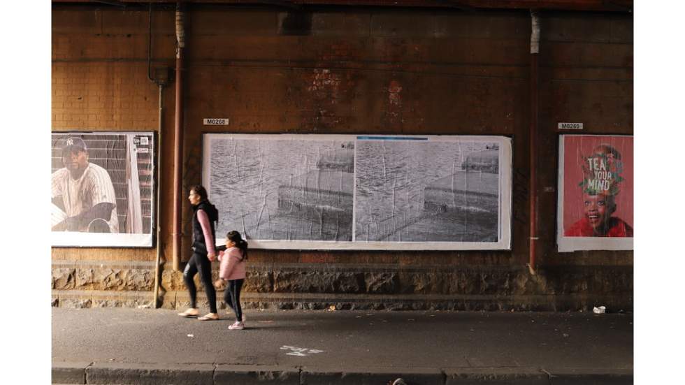 A woman and child holding hands walking through a bridge underpass. Large printed posters are fixed to the brick wall.  