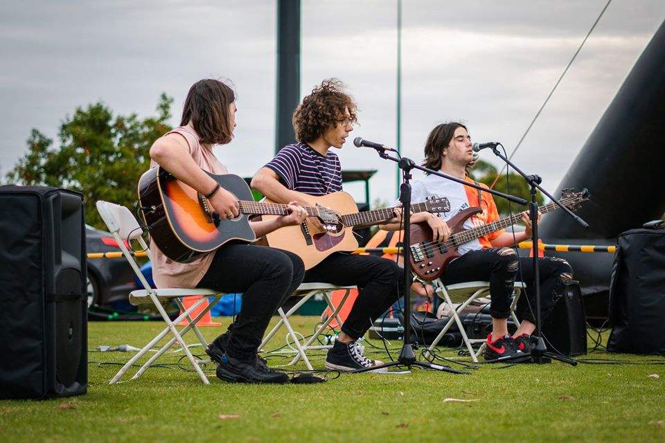 Three teenage boys seated and performing at an outside event playing acoustic guitar.