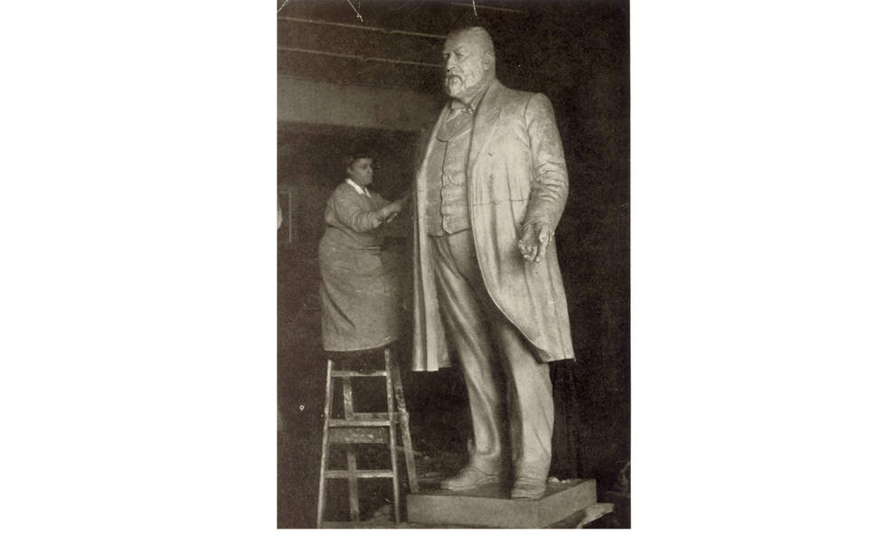 Black and white photo of a woman in an art smock standing on a ladder working on a larger than life sculpture of a man.