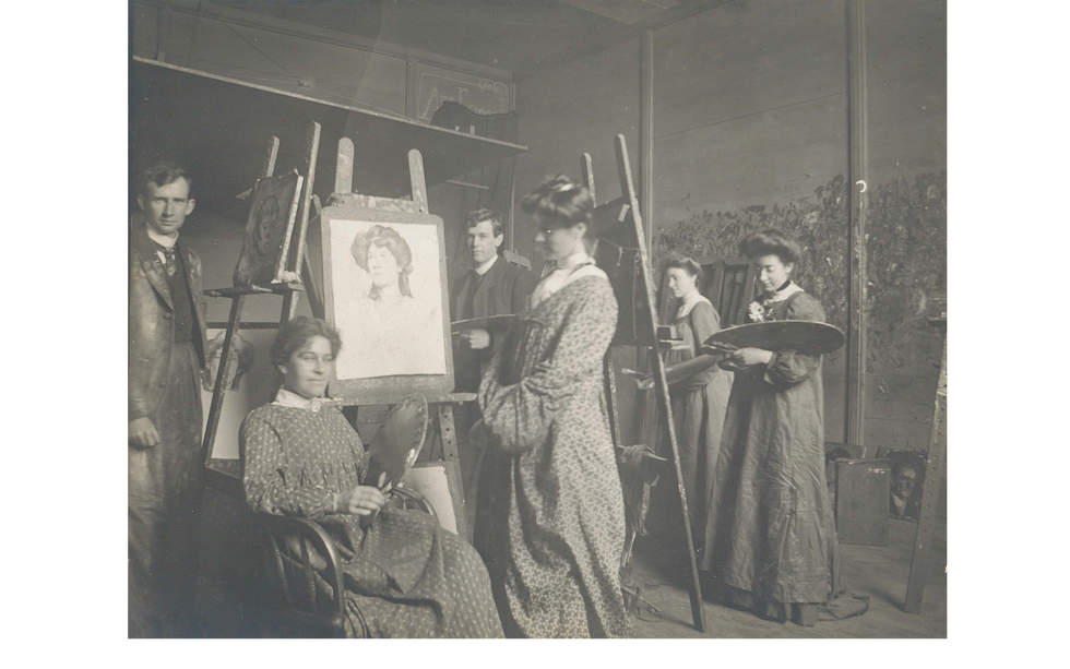 Black and white photo of an art studio with women and men painting holding easels and wearing art smocks.