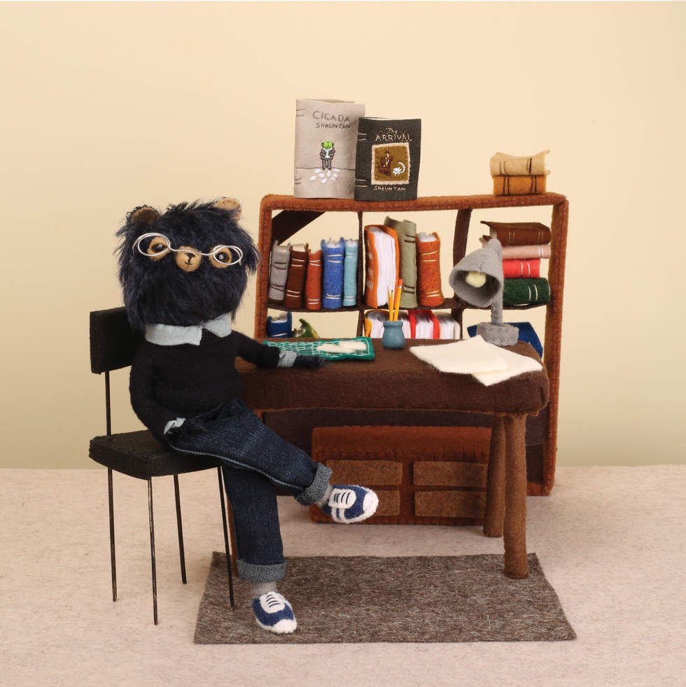 A three dimensional scene, made from felt and fabric, of a bear dressed as a person wearing glasses, seated at a desk with books in the background.