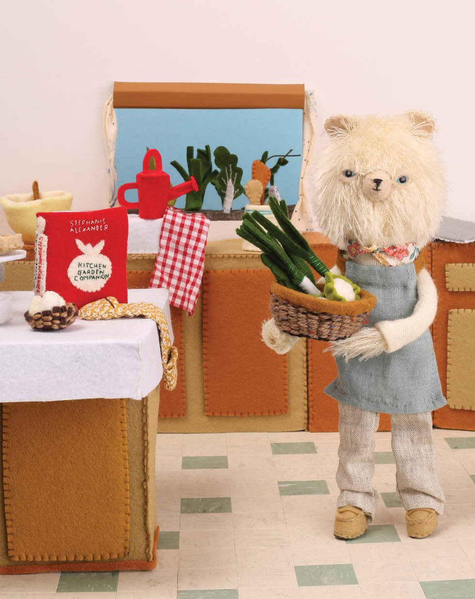 A three dimensional felted and fabric scene of a bear standing in a kitchen holding a basket of vegetables.