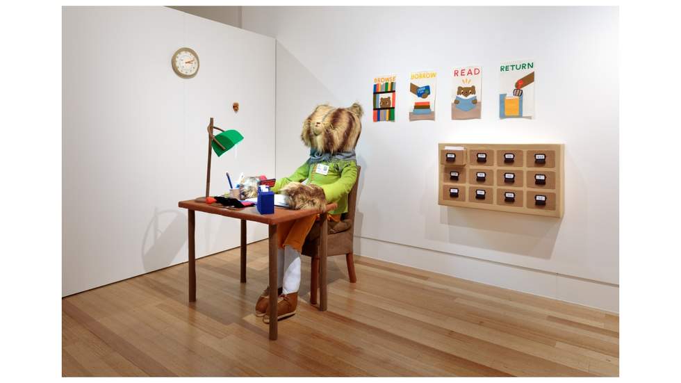 Interior view of an exhibition with a large bear seated at a desk.