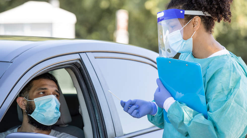 Health worker in full PPE with clipboard and swab talks to a man in a car