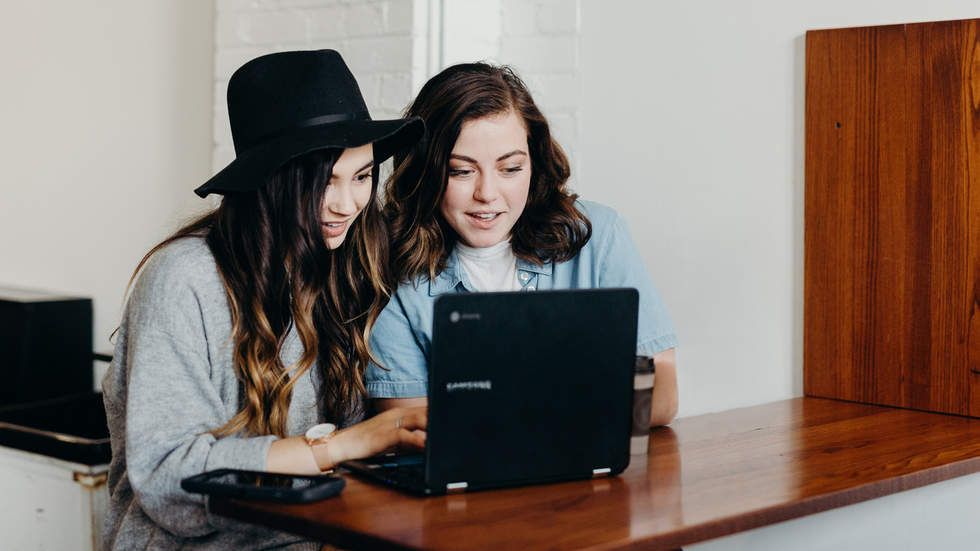Two young women looking at black laptop on bench