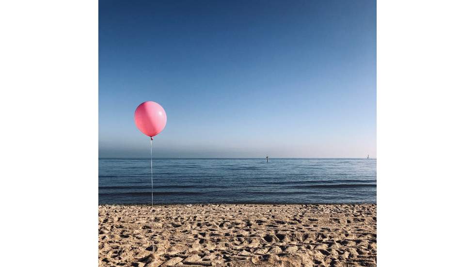 Photograph of an upright, pink balloon with string on a foreshore overlooking a calm flat sea and blue skyline.. 