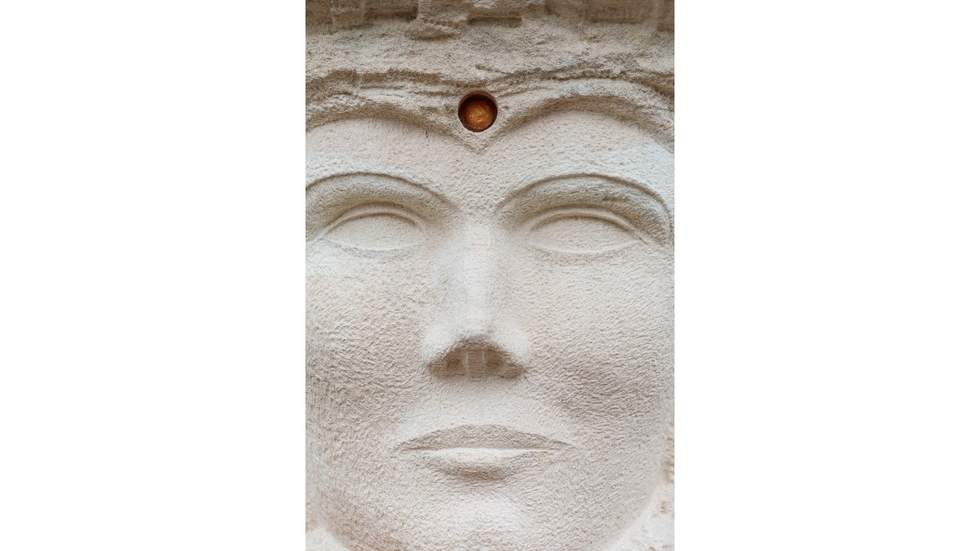 A close up view of a face made from limestone with a stone embedded in the center of the forehead. 