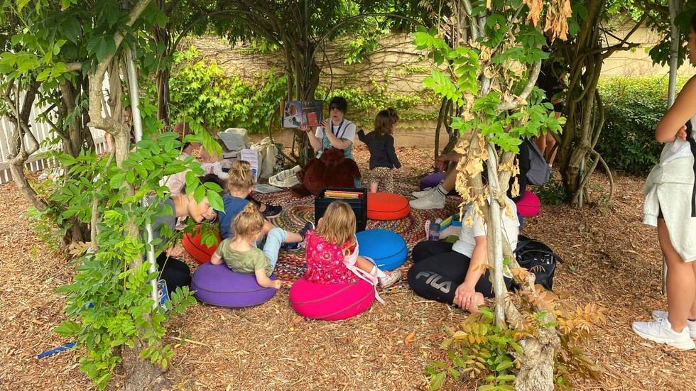 Lady reading story to kids sat on cushions under a tree 