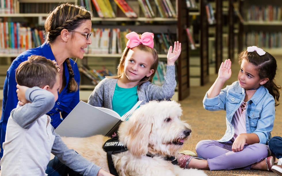 Lady reading book to children in library with a dog