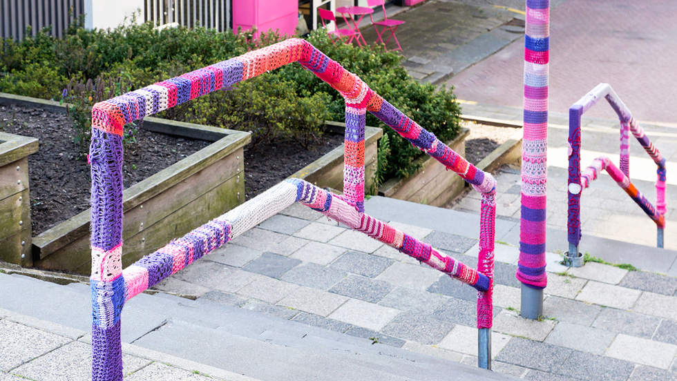 Pink and purple yarn bombing on poles and hand rails on a set of stairs with yarn.
