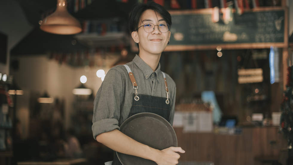 Photograph of a young man holding a tray in a cafe, he is smiling, and looks like he has just started a new job.