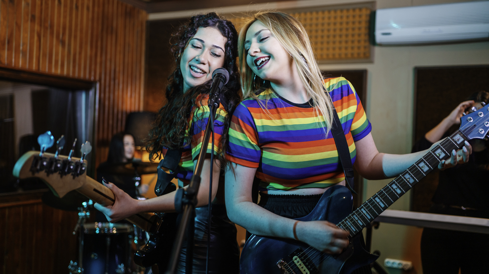 photo of two young people singing into a microphone. Left has long brown hair, rainbow t shirt and is holding a guitar. right young person has long blonde hair and is wearing a rainbow striped t shirt and playing a steel grey guitar. The microphone is between them.