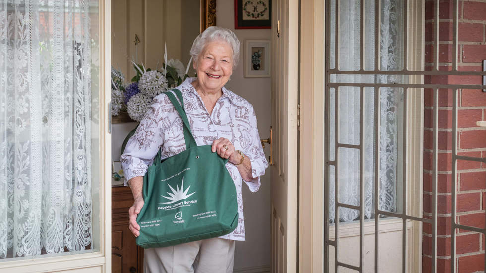 Older woman receiving her home library service delivery. She is standing at her front door holding the book bag and smiling.