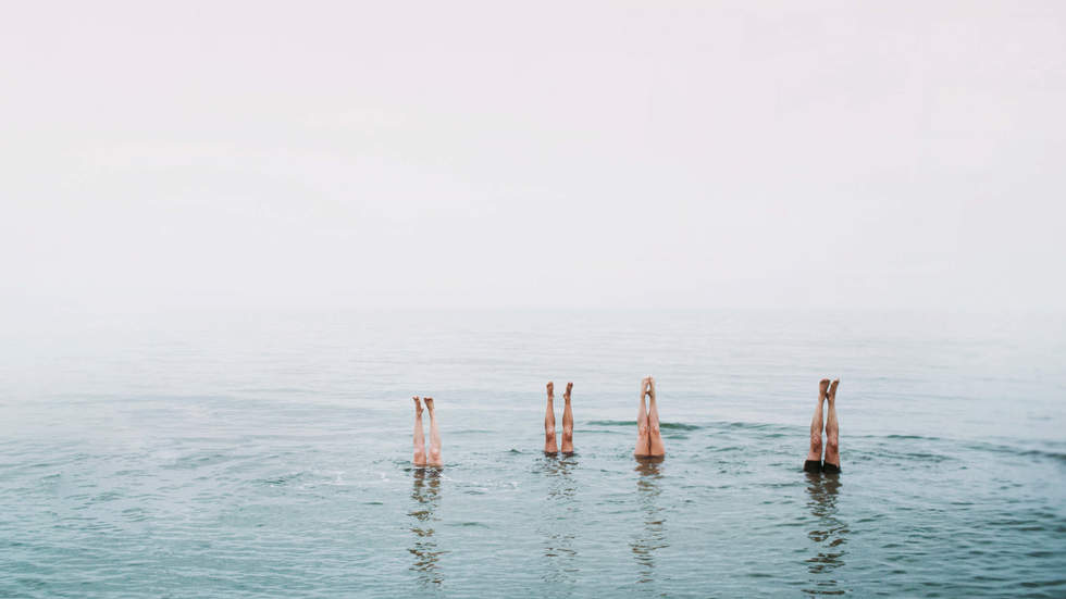 Bron Smart, Handstands, 2002. Four people doing handstands in the bay. The tops of their legs are showing above the water and the tones are soft pinks and blues.