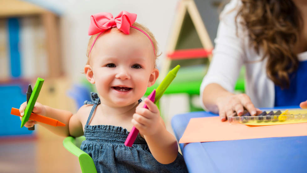 Little baby girl with a pink bow in her hair sitting at a table and holding crayons and smiling at the camera.