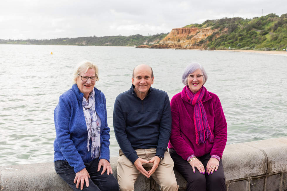 3 older people smiling at the camera, water behind them 