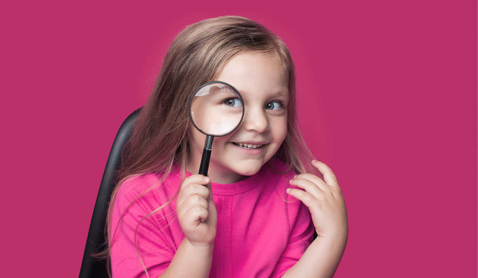 Great Art Detective - little girl holding a magnifying glass up to her eye and smiling. She is wearing a bright pink top and is against a pink background.
