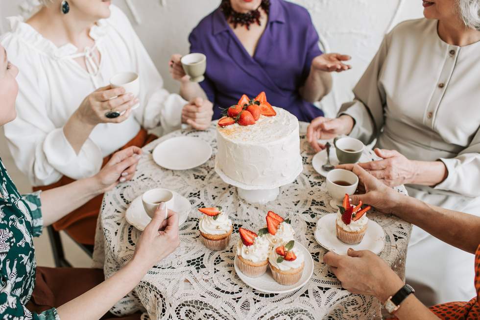 White cake with strawberries on top and people sitting around drinking tea. 