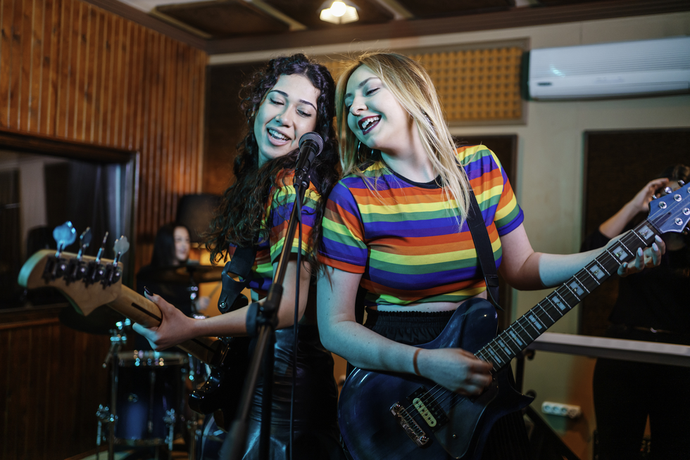 photo of two young people singing into a microphone. Left has long brown hair, rainbow t shirt and is holding a guitar. right young person has long blonde hair and is wearing a rainbow striped t shirt and playing a steel grey guitar. The microphone is between them.