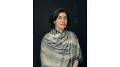 Photographed portrait of a woman, with black hair, seated in front of a black background wrapped in a printed grey and white textile.