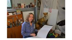Michelle Zuccolo sitting in her studio holding pencil and open sketch book with a taxidermy fox and books in the background.