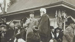 Black and white photograph of two men, one in a bowler hat, one with a grey beard, surrounded by people.