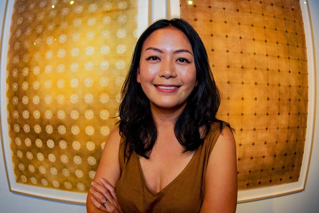 Catherine Pickop smiles at the camera in front of two framed artworks of circular geometrical forms of golden hues. 