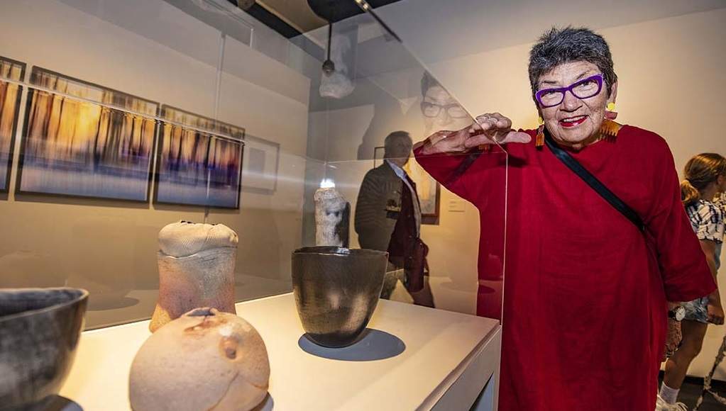 Jill Symes smiles towards the camera leaning on a glass display case with ceramic vessels displayed inside. 