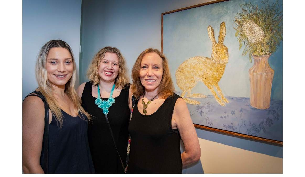 Three women smiling towards the camera standing next to a painting of a hare with vase of native flowers.   