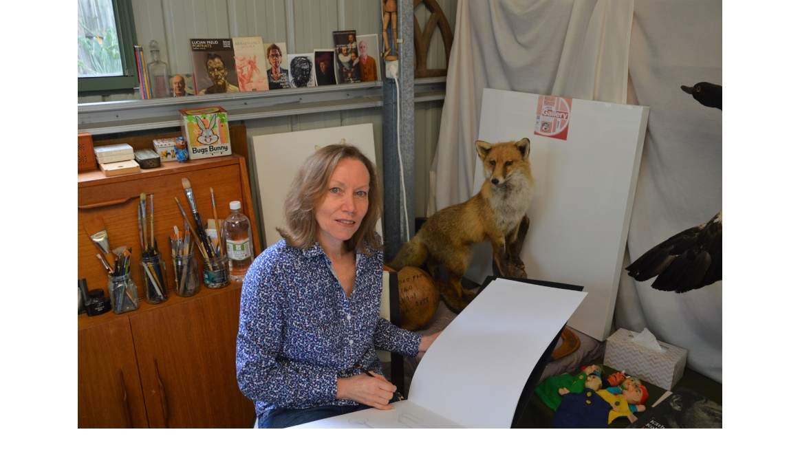 Michelle Zuccolo sitting in her studio holding pencil and open sketch book with a taxidermy fox and books in the background.