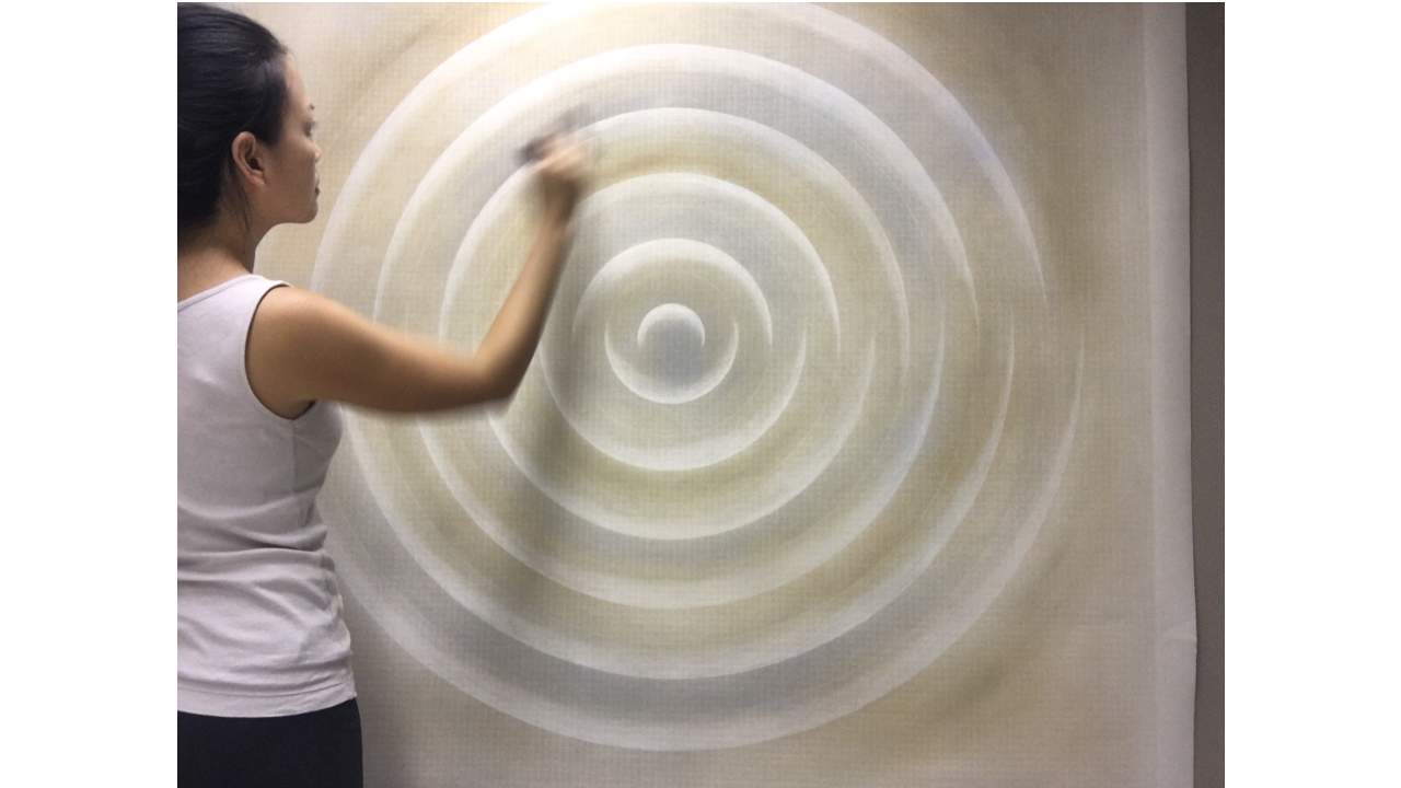 A woman standing drawing a spiral in shades of white, beige and grey on a large sheet of white paper. 