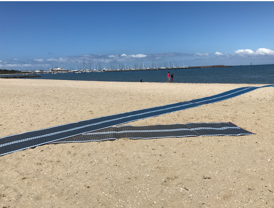 Access matting on Hampton Beach and two people walking at edge of water with boats in background 