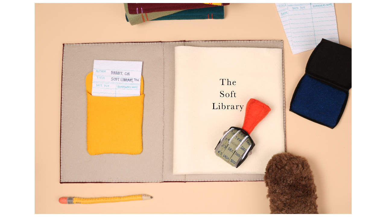 A book made from felt, with an open page showing the text ‘the soft library’ and a library stamp and borrowing card in the front pocket of the book.