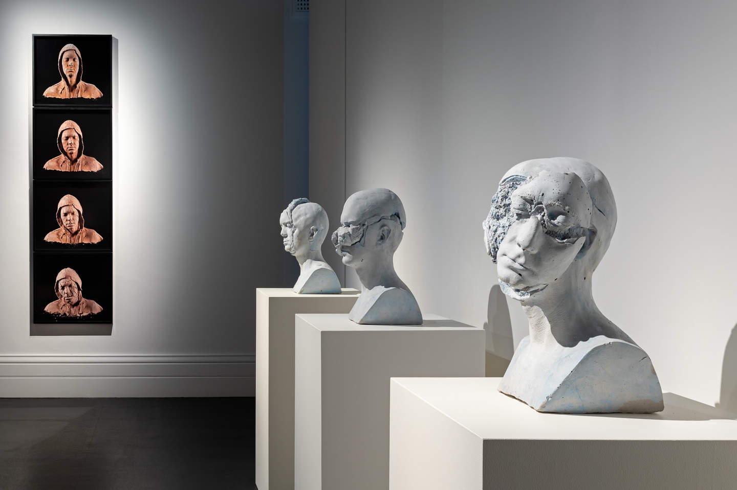 A gallery interior with sculpture busts on plinths and photographs of terracotta heads hung on wall.