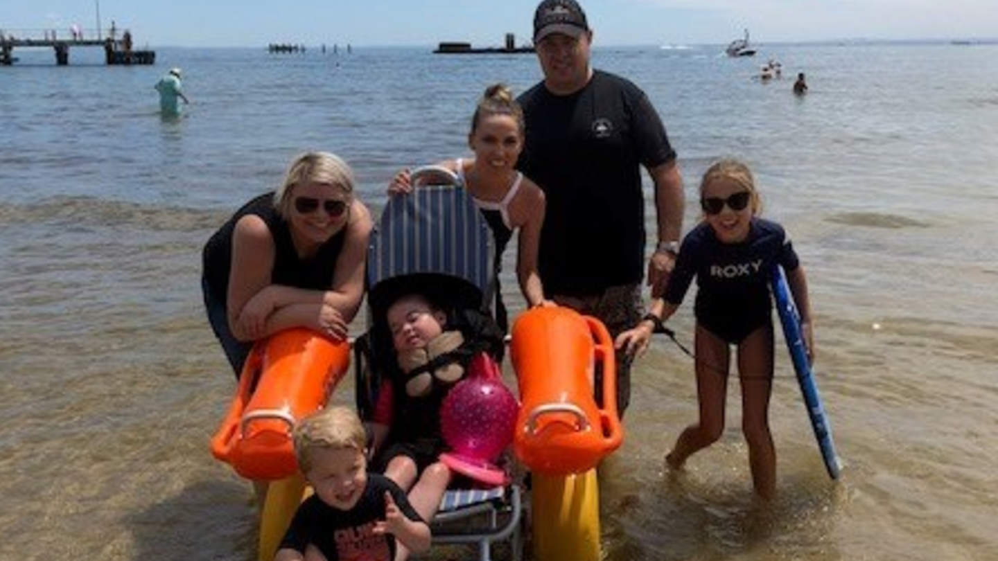 Young family and carer in shallow water with young child with a disability laying in chair 