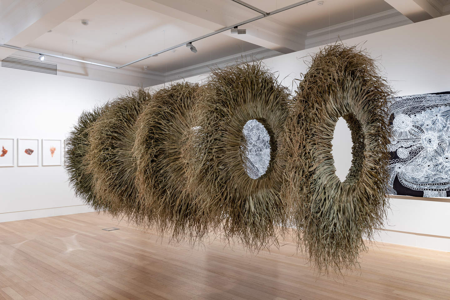 Five suspended circular wreaths made from green organic matter resembling long grass, aligned to create a cylinder.  