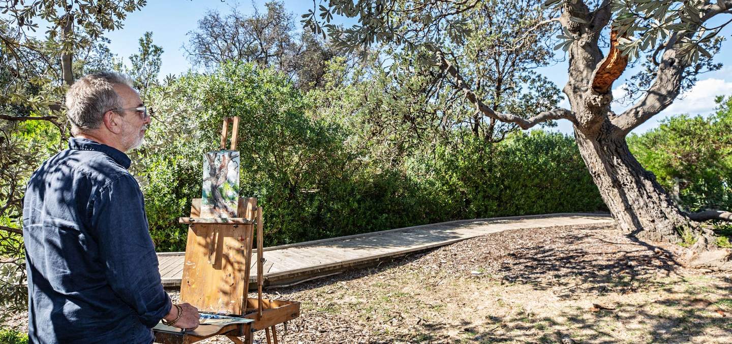 Gregory Alexander stands at an easel with a painting of a tree. He is surrounded by vegetation, the tree he paints is in front of him to the right.