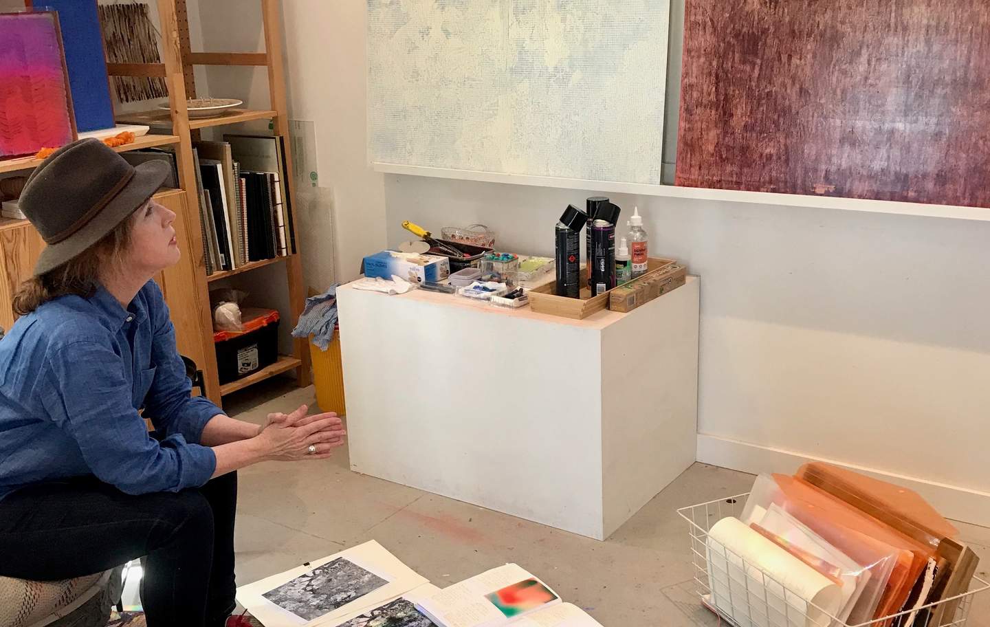 Kate Stewart seated in her art studio wearing an Akubra, looks thoughtfully up towards two paintings. 
