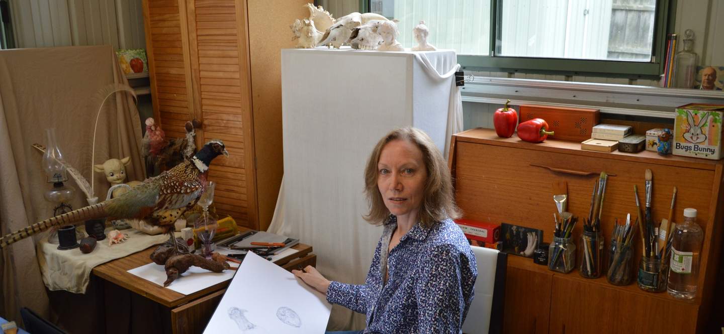 Michelle Zuccolo sits in her studio holding pencil and open sketch book with a taxidermy fox and books in the background