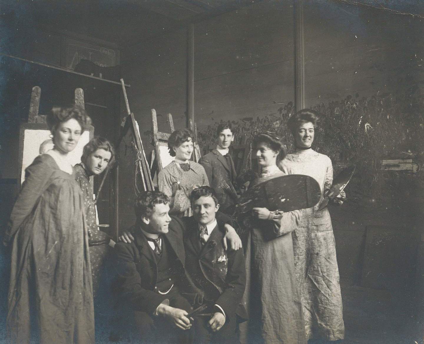 Black and white photograph of women and men student artists seated and standing, wearing art smocks, some holding painting palettes, with easels in the background.