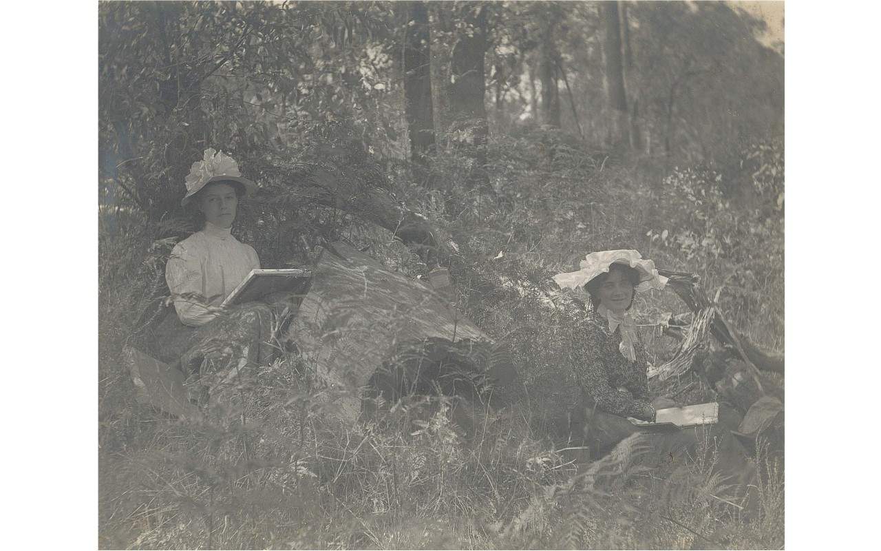 Black and white photograph of two woman in early 20th century clothing seated painting outdoors in tall grass.