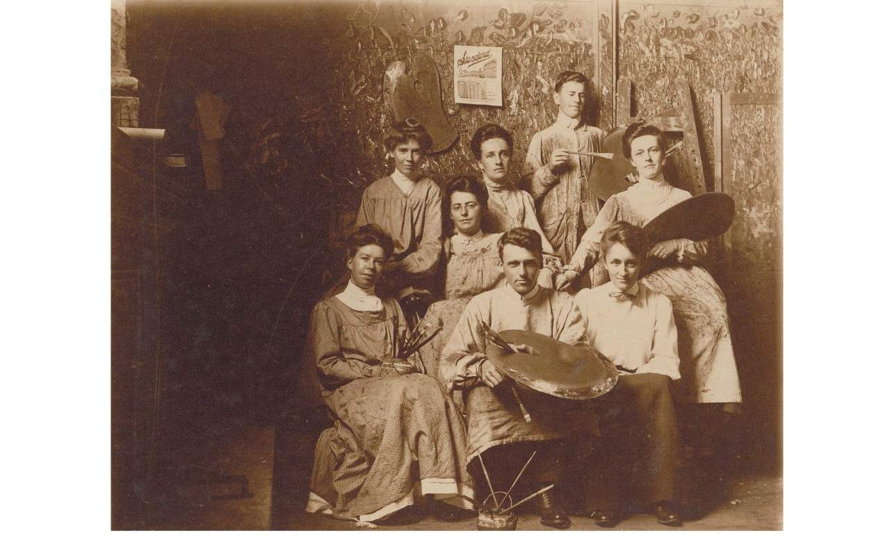 Black and white photo of men and women students seated and standing wearing smocks.
