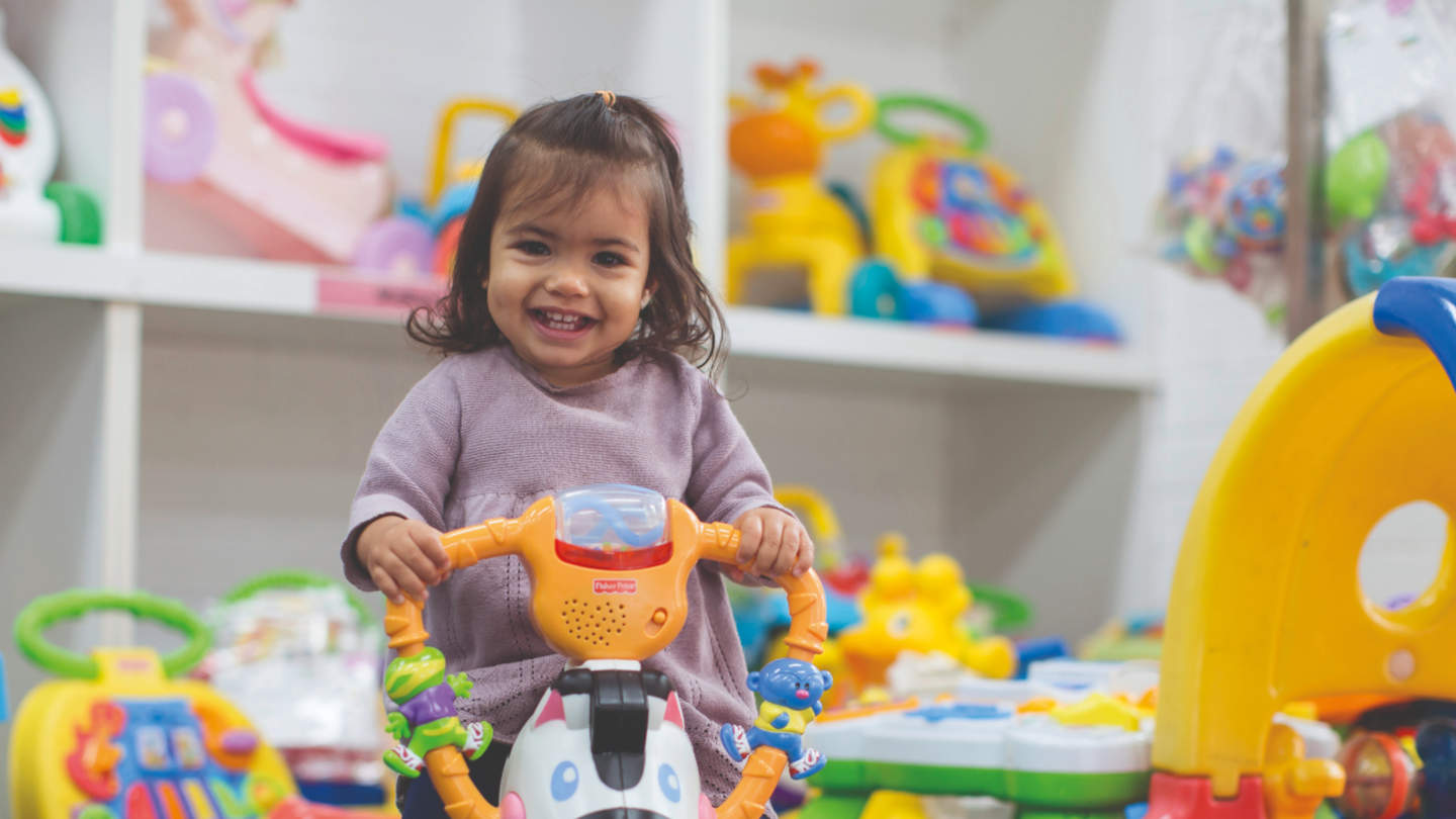 Young girl with brown hair smiling and holding onto toddler walker at Toy Library