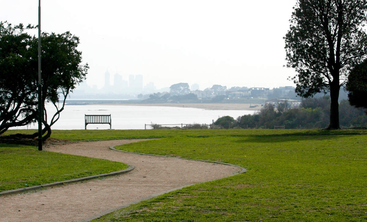 Pathway leading through grass and to a foreshore bench overlooking Port Phillip Bay and Melbourne CBD skyline.