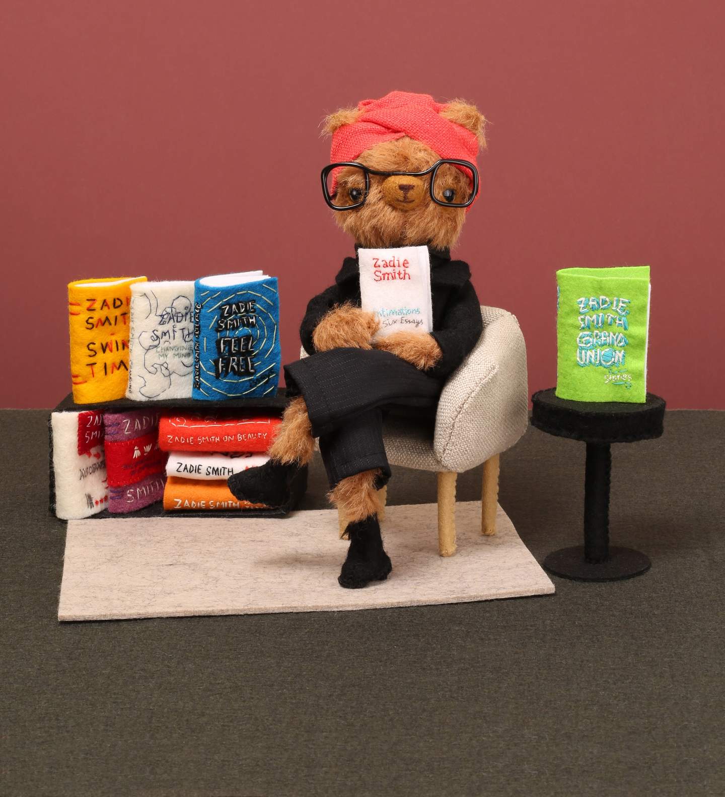 A three dimensional felted and fabric scene of books on a shelf and a seated bear holding up a book.