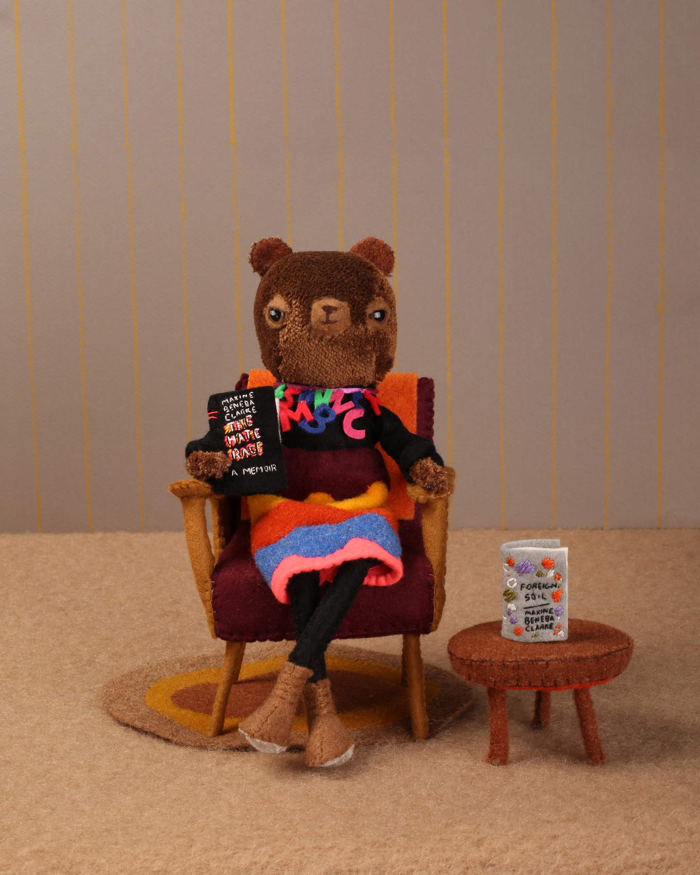 A bear dressed in a colourful dress seated in a lounge chair holding up a book