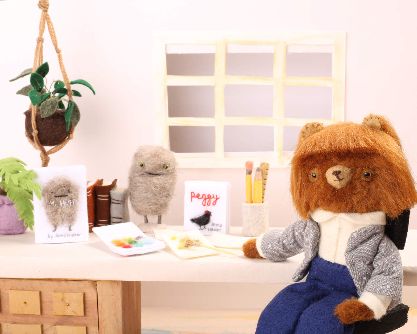 A 3D felted scene of a bear in clothes seated at a desk with books and toy.