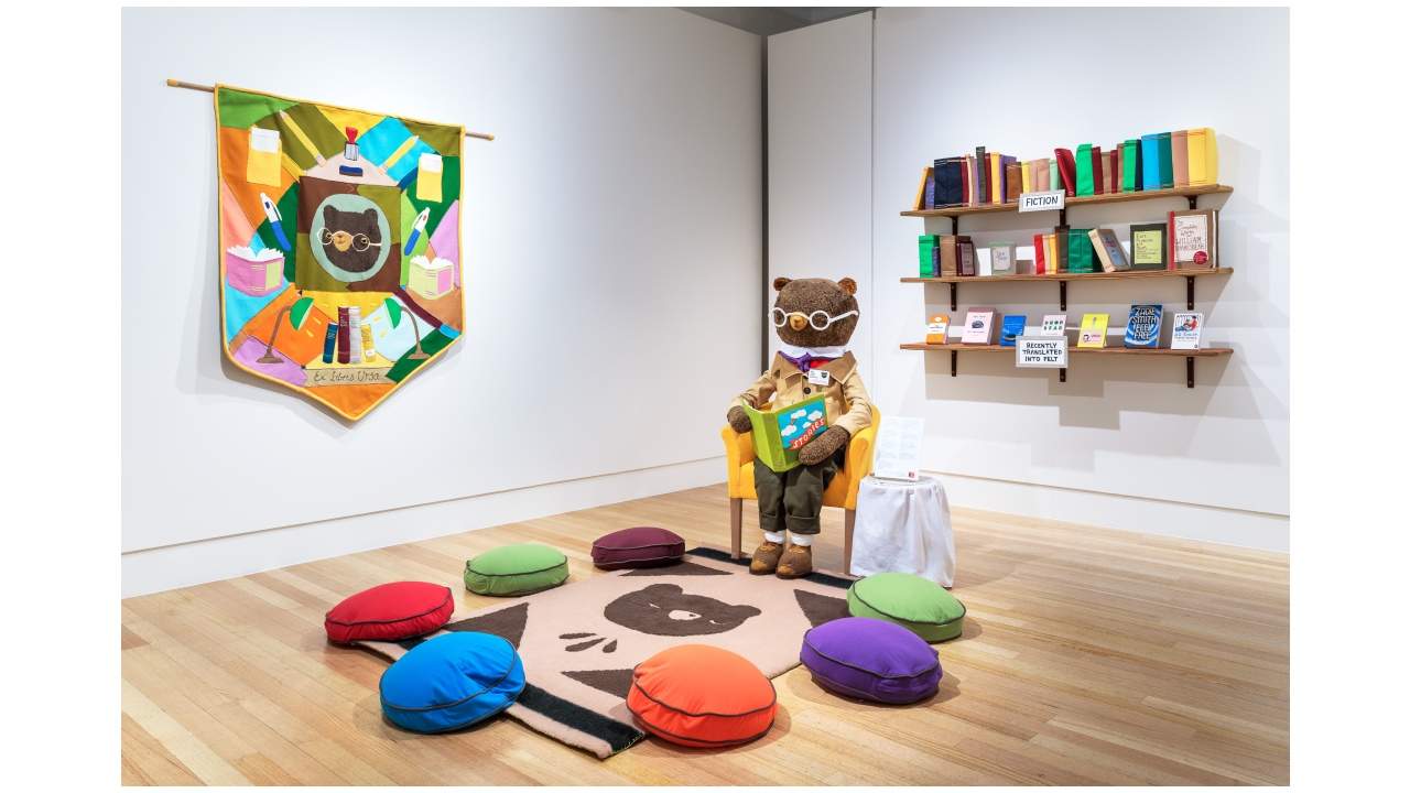 Interior view of an exhibition with a large bear seated on chair holding a book.