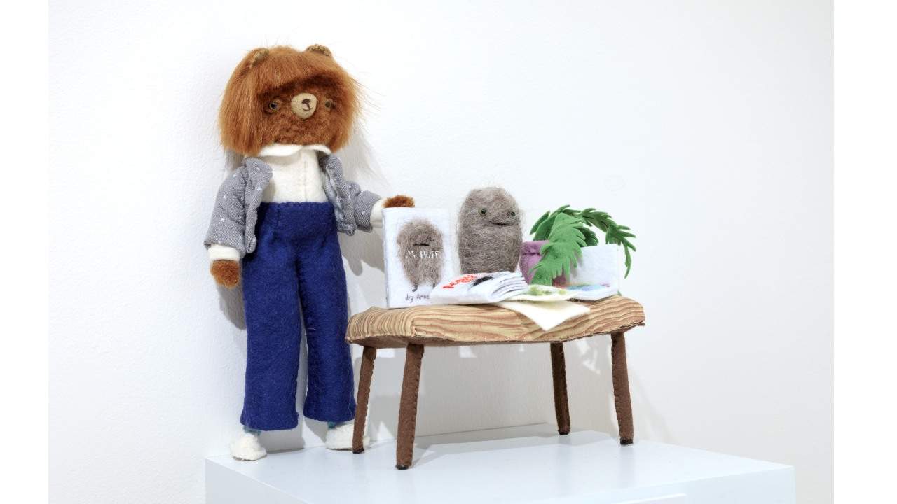 A 3D scene, made from felt, of a bear dressed as a person standing next to a table with a book and plant.